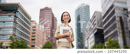 Portrait of young asian woman, looking happy and confident, going to work or university, city skyscrappers behind her, holding laptop and notebook Portrait of young asian woman, looking happy and confident, going to work or university, city skyscrappers behind her, holding laptop and notebook 107443295