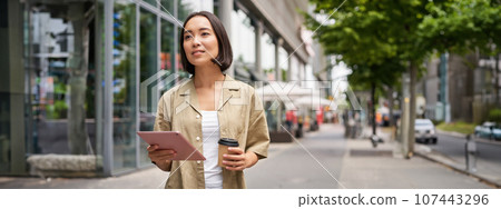 Portrait of smiling young woman walking in city with tablet, drinking takeaway coffee, going down the street with happy expression Portrait of smiling young woman walking in city with tablet, drinking takeaway coffee, going down the street with happy expression 107443296