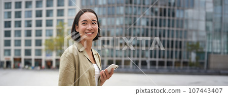 Vertical shot of young asian digital nomad, girl talks on mobile phone and walks on street with laptop. Young woman remote worker going to coworking space 107443407