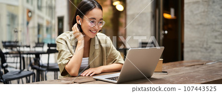 Portrait of young woman video chatting, having online meeting on laptop, sitting in outdoor cafe with compute, wearing glasses and smiling Portrait of young woman video chatting, having online meeting on laptop, sitting in outdoor cafe with compute, wearing glasses and smiling 107443525