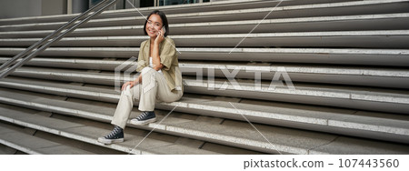 Smiling asian girl sits on stairs of building and talks on mobile phone, relaxing during telephone conversation Smiling asian girl sits on stairs of building and talks on mobile phone, relaxing during telephone conversation 107443560