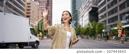 Happy asian girl passing by friend and waving at them on street, saying hello while walking in city, holding smartphone 107443643
