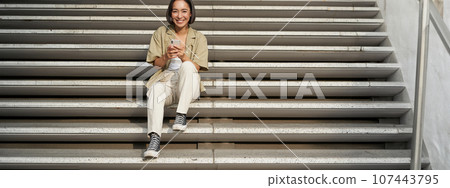 Portrait of smiling asian girl sits on stairs with her smartphone, browsing internet on mobile phone, resting outdoors in city centre Portrait of smiling asian girl sits on stairs with her smartphone, browsing internet on mobile phone, resting outdoors in city centre 107443795