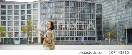 Modern people. Young woman walks on street of city and listens music in wireless headphones, holds smartphone Modern people. Young woman walks on street of city and listens music in wireless headphones, holds smartphone 107443810