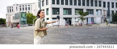 Car sharing and technology. Young asian woman waiting for taxi near road, holding smartphone, order car in application 107443811