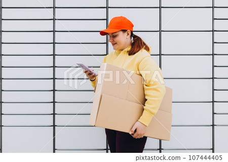 Young woman in red hat picks up parcel from automatic post office machine, Courier standing with phone and small box. Concept of fast delivery to automatic self lockers 107444405