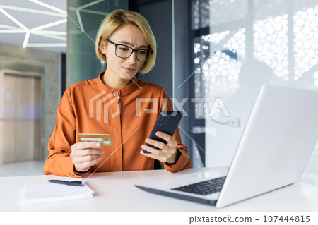 Upset and disappointed woman inside office holding bank credit card and phone, business woman received a fund transfer rejection error, female worker at workplace with laptop. Upset and disappointed woman inside office holding bank credit card and phone, business woman received a fund transfer rejection error, female worker at workplace with laptop. 107444815