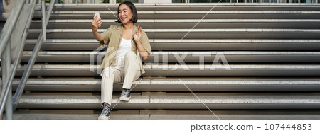 Portrait of asian beautiful girl takes selfie on smartphone. Young korean woman sits on stairs outdoors and makes photos on mobile phone Portrait of asian beautiful girl takes selfie on smartphone. Young korean woman sits on stairs outdoors and makes photos on mobile phone 107444853