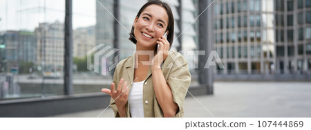 Cellular connection. Young asian woman makes a telephone call, talking on mobile smartphone and walking on street Cellular connection. Young asian woman makes a telephone call, talking on mobile smartphone and walking on street 107444869