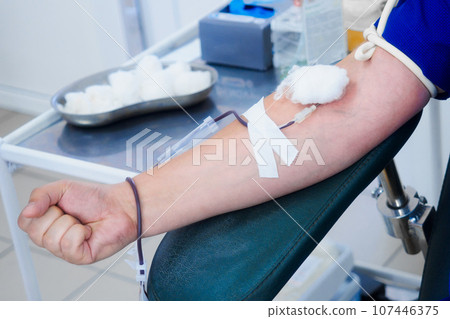Donor sits in chair and donates blood from vein in medical clinic. Donor's hand with IV and needle 107446375