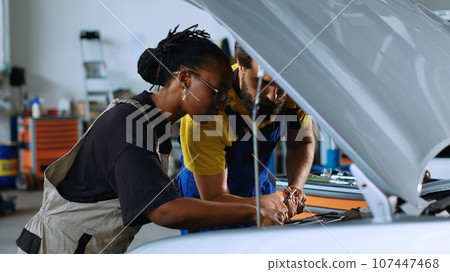 Coworking technicians in car service using torque wrench to fix damaged automobile. Man and woman in repair shop servicing broken vehicle, checking for faulty transmission using professional tools 107447468