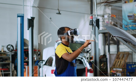African american mechanic in car service using virtual reality goggles to visualize automobile components in order to fix them. Repairman wearing futuristic vr headset to test vehicle engine African american mechanic in car service using virtual reality goggles to visualize automobile components in order to fix them. Repairman wearing futuristic vr headset to test vehicle engine 107447477