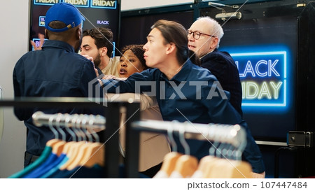 Security officer using walkie talkie, handling crowd control on black friday big sales event. Aggressive shoppers trying to break red tape to enter retail store boutique, clearance items. 107447484