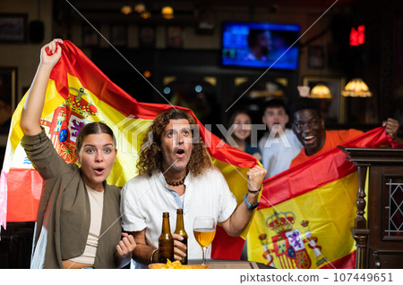 Excited diverse sport fans with Spanish flag rejoicing winning game with glasses of beer and chips in the pub 107449651