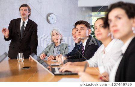 Displeased male manager scolding at young, adult, mature office workers sitting with reports and laptops at table in meeting room Displeased male manager scolding at young, adult, mature office workers sitting with reports and laptops at table in meeting room 107449739