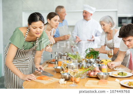 Young Asian woman cutting fresh salmon during group cooking lesson Young Asian woman cutting fresh salmon during group cooking lesson 107449768