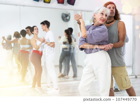 Happy caucasian guy and old lady practising paired latin dance in dance studio Happy caucasian guy and old lady practising paired latin dance in dance studio 107449770