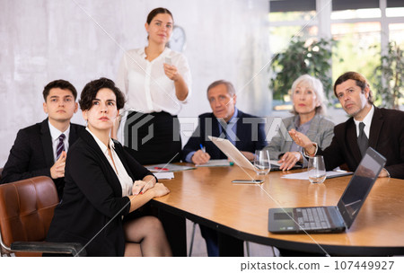 Colleagues of different ages using laptops and watching presentation while young female turning on presentation in office Colleagues of different ages using laptops and watching presentation while young female turning on presentation in office 107449927