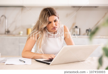 Disappointed woman sitting at table and looking at display of laptop 107450003