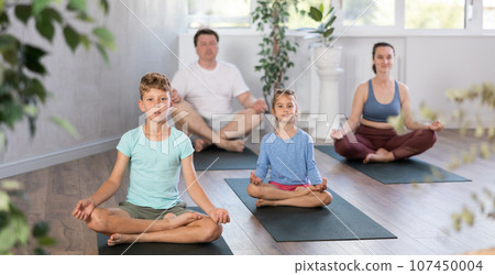 Positive pre-teen girl performing yoga exercises with brother and parents at gym 107450004