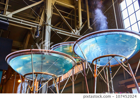 Giant vats of water in clear bowls on long copper stands with one steaming and spitting water 107452024