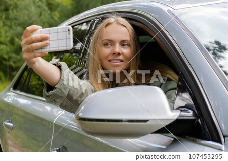 Exited smiling woman making video call with mobile phone from car window. Local solo travel on weekends concept. Young traveler explore freedom outdoors in forest taking selfie photo. Unity with 107453295
