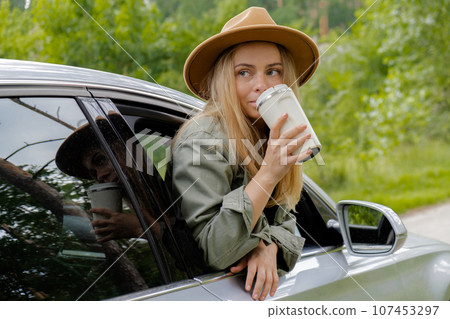 Blonde woman sticking head out of windshield car and drink coffee or tea from reusable mug. Young tourist explore local travel making candid real moments. Refuse reuse recycle zero waste concept Blonde woman sticking head out of windshield car and drink coffee or tea from reusable mug. Young tourist explore local travel making candid real moments. Refuse reuse recycle zero waste concept 107453297