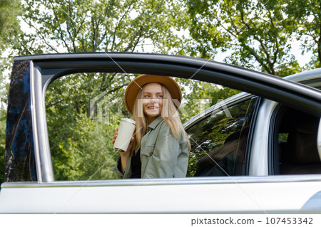 Blonde woman stoped on road next to car and drink coffee or tea from reusable mug. Refuse reuse recycle zero waste concept. Young tourist explore local travel making candid real moments. Responsible Blonde woman stoped on road next to car and drink coffee or tea from reusable mug. Refuse reuse recycle zero waste concept. Young tourist explore local travel making candid real moments. Responsible 107453342