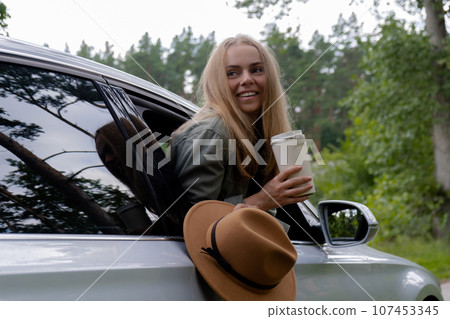 Smiling young woman looking from car window and drinking coffee or tea from reusable thermos cup. Local solo travel on weekends concept. Exited woman explore freedom outdoors in forest. Unity with Smiling young woman looking from car window and drinking coffee or tea from reusable thermos cup. Local solo travel on weekends concept. Exited woman explore freedom outdoors in forest. Unity with 107453345