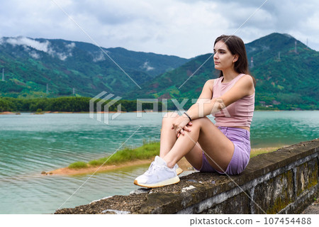Young russian woman sitting with depressed and sad face against lake and mountains in Khanh Hoa Province, Vietnam Young russian woman sitting with depressed and sad face against lake and mountains in Khanh Hoa Province, Vietnam 107454488