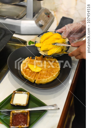 the cook putting fresh sliced mango fruit on baked belgian waffles with tongs the cook putting fresh sliced mango fruit on baked belgian waffles with tongs 107454516
