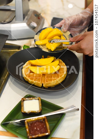 the cook putting fresh sliced mango fruit on baked belgian waffles with tongs the cook putting fresh sliced mango fruit on baked belgian waffles with tongs 107454517