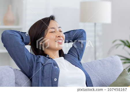 Dreamy asian woman relaxing at home close up, woman smiling and thinking sitting on sofa with hands behind head with closed eyes. Dreamy asian woman relaxing at home close up, woman smiling and thinking sitting on sofa with hands behind head with closed eyes. 107458167
