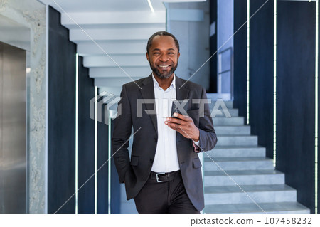 Portrait of happy successful African American boss, man smiling and looking at camera, businessman holding phone using online application, man walking down corridor of building inside office. Portrait of happy successful African American boss, man smiling and looking at camera, businessman holding phone using online application, man walking down corridor of building inside office. 107458232