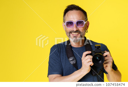 Happy, enthusiastic photographer smiles with delight while holding mirrorless camera against yellow background. Joy and passion that photographer feels when capturing moments with beloved camera. High 107458474