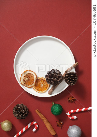 Creative background with christmas holiday concept for advertising. On the red background, white round dish decorated with dry pine cone, cinnamon stick, candy cane and colorful baubles Creative background with christmas holiday concept for advertising. On the red background, white round dish decorated with dry pine cone, cinnamon stick, candy cane and colorful baubles 107460691
