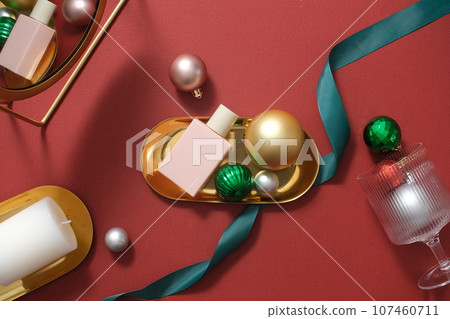 Scene for advertising and branding product with Christmas concept. A pink cosmetic bottle placed on golden tray with decorations: baubles, ribbons and candles on red background Scene for advertising and branding product with Christmas concept. A pink cosmetic bottle placed on golden tray with decorations: baubles, ribbons and candles on red background 107460711