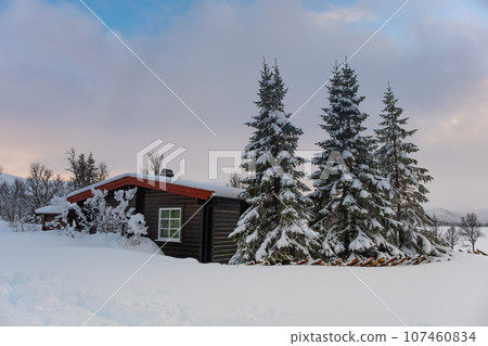A small snow covered hunting lodge in the norwegian mountains with animal traces in the foreground and mountain summits in the background A small snow covered hunting lodge in the norwegian mountains with animal traces in the foreground and mountain summits in the background 107460834
