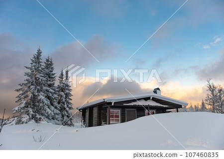 A small snow covered hunting lodge in the norwegian mountains with animal traces in the foreground and mountain summits in the background 107460835