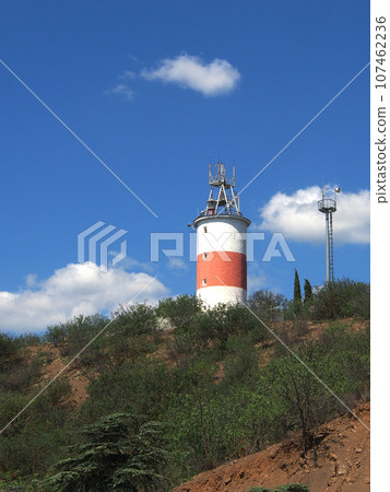 A lighthouse on a steep, rocky shore overgrown with sparse vegetation, against a blue sky. Hot summer day, travel, tourism. 107462236