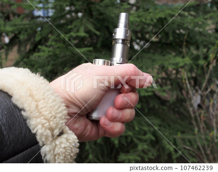 VAPE concept, a man's hand shows a vape device on a blurry green background. The concept of vaping. VAPE shop. Smoking electronic cigarettes. Sale of electronic cigarettes. VAPE concept, a man's hand shows a vape device on a blurry green background. The concept of vaping. VAPE shop. Smoking electronic cigarettes. Sale of electronic cigarettes. 107462239