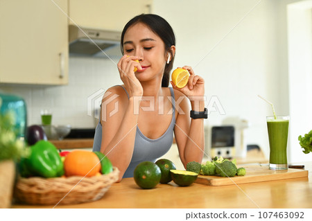 Beautiful young woman smelling fresh lemon with closed eyes while preparing ingredients for making healthy food in kitchen Beautiful young woman smelling fresh lemon with closed eyes while preparing ingredients for making healthy food in kitchen 107463092