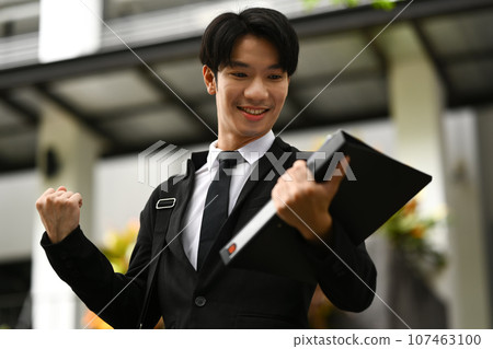 Handsome businessman celebrating success, clenching fists while standing outside business center 107463100