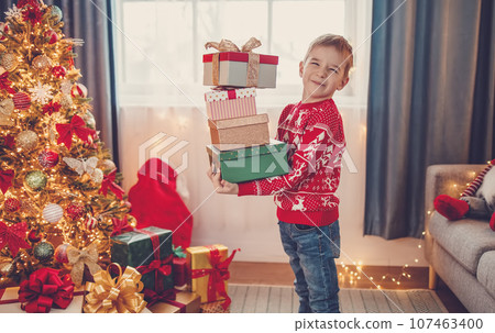 Boy standing near Christmas tree with stack of gift boxes in his hands. 107463400