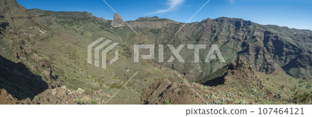 Panoramic scenic landscape with Roque de Agando rock at hiking trail through Barranco de Guarimiar Gorge. Green mountain canyon walls, blue sky background. Copy space. La Gomera, Canary Islands, Spain Panoramic scenic landscape with Roque de Agando rock at hiking trail through Barranco de Guarimiar Gorge. Green mountain canyon walls, blue sky background. Copy space. La Gomera, Canary Islands, Spain 107464121