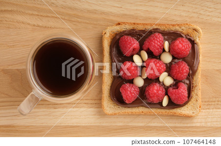 A cup of coffee and a sandwich with chocolate paste and raspberries on a wooden background. Top view. A cup of coffee and a sandwich with chocolate paste and raspberries on a wooden background. Top view. 107464148