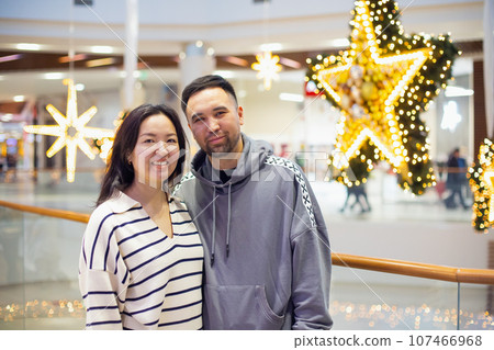 young Asian Kazakh male and female couple in shopping mall on Christmas or New Year holidays. happy family or man and woman in love against huge decor stars in public place. Valentine's Day, newlyweds 107466968