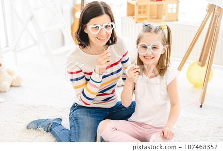 Cute little girl and mother holding stick glasses in kids room 107468352