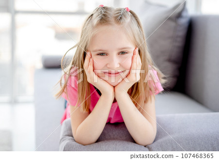 Happy little girl resting on sofa with head on hands indoors 107468365