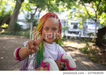 little cute girl with colorful afro pigtails sits on a skateboard and shows a peace sign 107468699